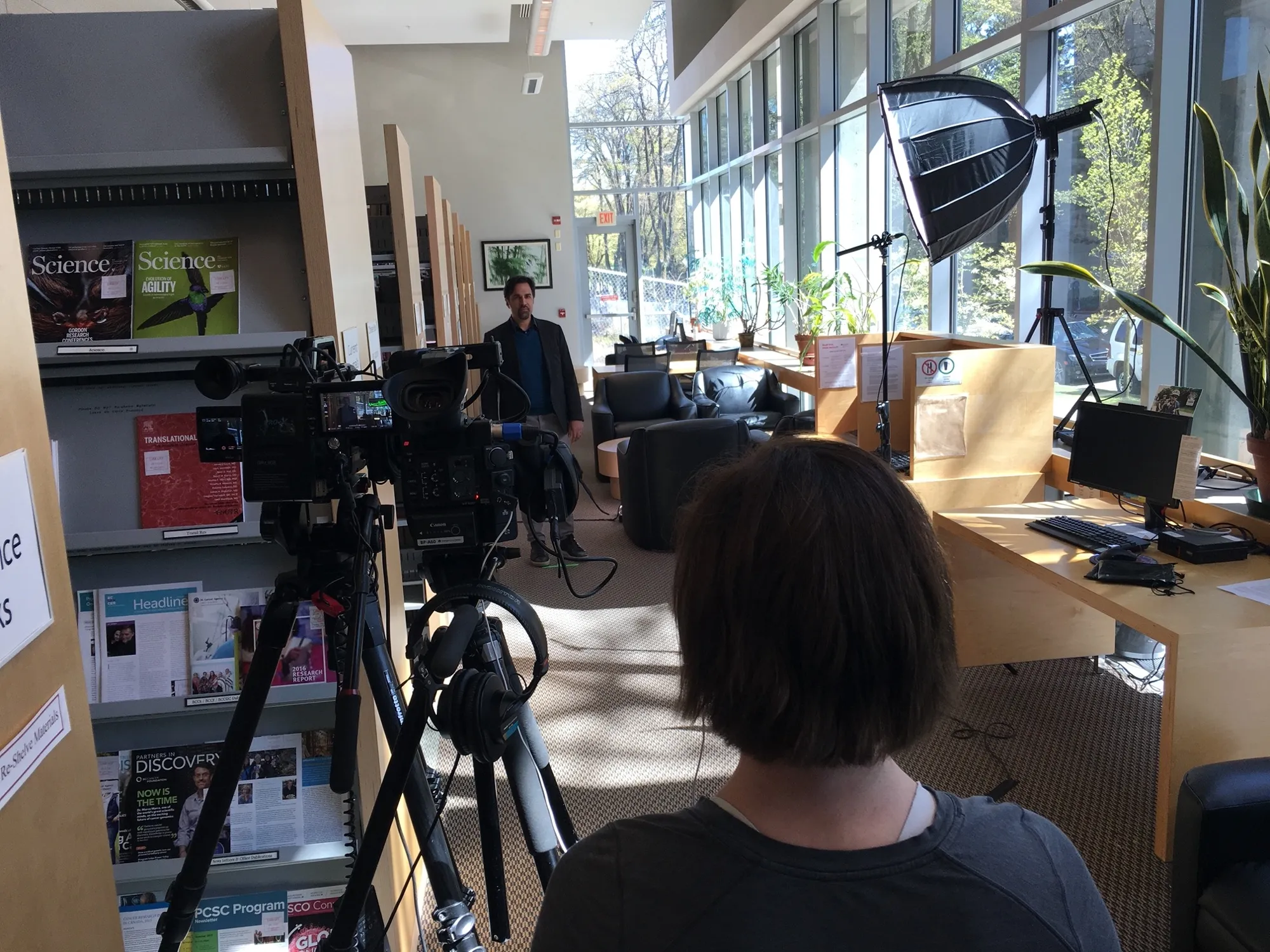 A cinema camera and boom rig set up to film an interview in the BC Cancer research library, the subject standing by shelves of Science journals.