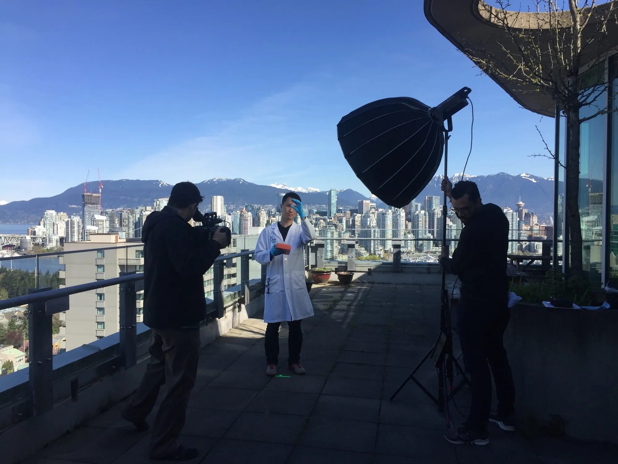 A rooftop shoot above the Vancouver skyline: a researcher in a white coat holds a sample as the crew films with a softbox key light.